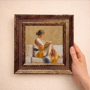 May include: A square framed painting depicting a person with dark hair tied back, sitting on a light-coloured sofa while reading an open book. The person wears a draped garment with yellow, white, and muted red tones. The frame is dark, textured gold.