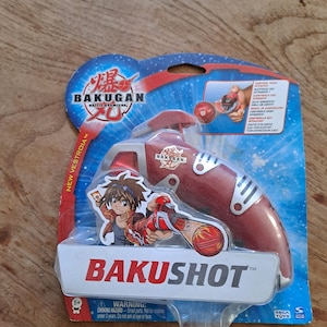 Op de afbeelding: Een rood en zilver Bakugan Bakushot speelgoed op een blauw-witte kaart. De verpakking toont het Bakugan-logo, een stripfiguur en de tekst "Bakushot". Het speelgoed is ontworpen voor kinderen van 5 jaar en ouder.