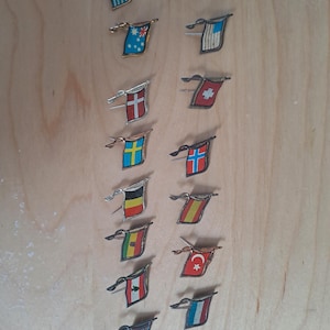 Op de afbeelding: Verzameling vintage vlagspelden, elk met een klein banierontwerp. De spelden tonen vlaggen van verschillende landen, waaronder Griekenland, Frankrijk en Australië. De spelden zijn gemaakt van metaal en hebben een speldrug.
