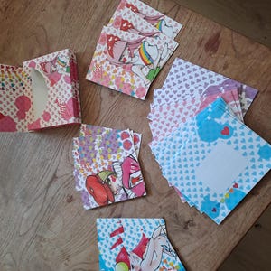 May include: A collection of colorful stationery items, including a box and several square cards, are arranged on a wooden surface. The cards feature illustrations of anime-style characters, hearts, and rainbows. The color palette includes pink, blue, and purple.