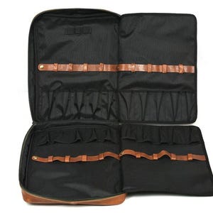May include: Open black travel case with multiple compartments and brown leather accents. The case has several pockets and leather straps for securing items. The case is open, revealing the interior organisation.