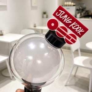 Op de afbeelding: Een heldere, bolvormige lichtstick met een zwarte basis en rode accenten. De lichtstick heeft de naam "JUNG KOOK" in witte letters op een rood rechthoekig stuk. De achtergrond toont een café met witte tafels en stoelen.