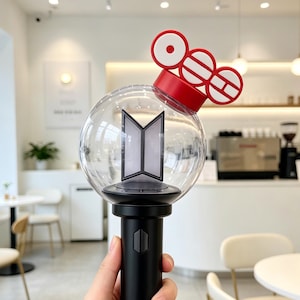 Puede incluir: Un bastón de luz negro y transparente con una parte superior roja. El bastón de luz tiene un globo transparente con un emblema gris en el interior. El mango es negro. El bastón de luz está sostenido por una mano.