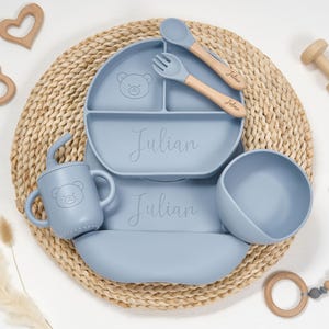 Könnte beinhalten: Ein hellblaues Baby-Ess-Set, bestehend aus einem geteilten Teller, einer Schüssel, einem Becher sowie Löffel und Gabel. Das Set hat ein Bären-Design und der Name "Julian" ist auf den Teller geschrieben. Holzspielzeug und eine Flechtmatte vervollständigen die Szene.