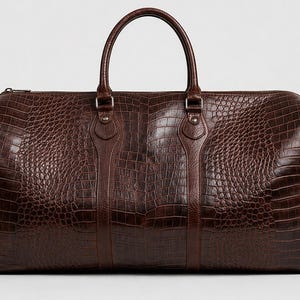 May include: A brown leather duffel bag with a crocodile skin texture. The bag has a top handle and a zip closure. The bag is a dark brown colour and appears to be made of high-quality leather. The bag is designed for travel.