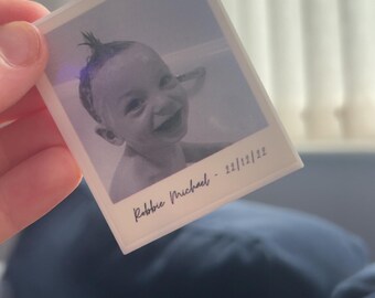 May include: A clear acrylic keychain featuring a grayscale photo of a smiling baby. The photo is in a Polaroid style with the name "Robbie Michael" and the date "22/12/22" printed at the bottom. The keychain has a silver-colored loop.