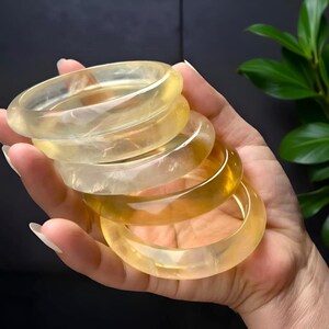 Könnte beinhalten: Eine Hand hält einen Stapel durchscheinender gelb-oranger Edelsteinarmreifen. Die Armreifen variieren in der Farbe von blassgelb bis zu einem tieferen Bernsteinton. Die glatten, polierten Oberflächen reflektieren das Licht.