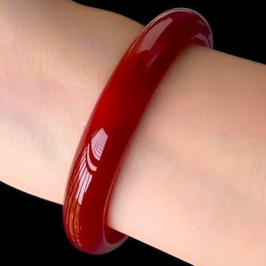 Puede incluir: Una pulsera tipo brazalete de color rojo sólido, lisa y con un acabado pulido y brillante. El accesorio circular presenta un perfil redondeado y un tono carmesí profundo y vibrante que refleja la luz sobre su superficie curva y uniforme.