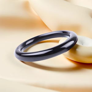 Può includere: Un braccialetto nero lucido. Il braccialetto circolare è realizzato in un materiale lucido. Poggia su una superficie color crema accanto a un piccolo oggetto rotondo color crema.
