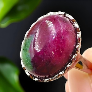 May include: A silver ring with a large, oval gemstone. The gemstone is a deep red with a green section. The ring has a decorative, textured border. The ring is a piece of jewellery.