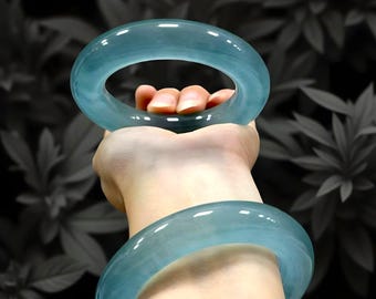 May include: Two translucent, light blue jade bangles are held in a hand against a blurred background of dark leaves. The bangles are smooth and circular, showcasing a subtle, natural color variation. The jade appears polished and reflects light.