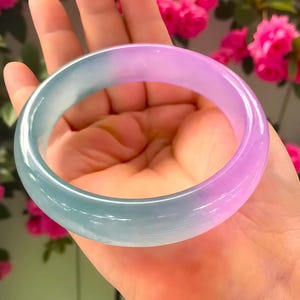 May include: A translucent jade bangle bracelet with a gradient of colors, transitioning from a light teal to a soft lavender. The smooth, polished surface reflects light, showcasing the natural variations in the stone. The bracelet is held in a hand, with a backdrop of pink flowers.