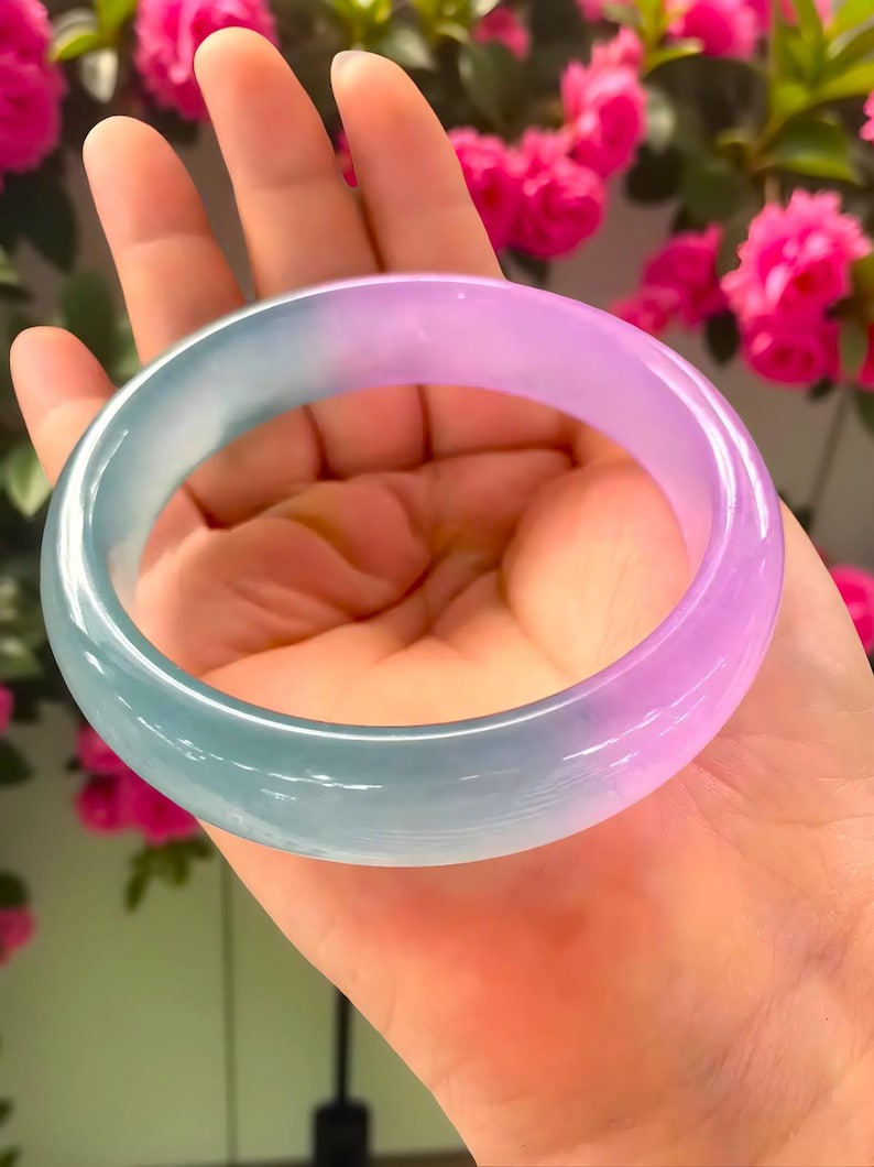 May include: A translucent jade bangle bracelet with a gradient of colors, transitioning from a light teal to a soft lavender. The smooth, polished surface reflects light, showcasing the natural variations in the stone. The bracelet is held in a hand, with a backdrop of pink flowers.