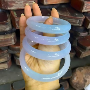 May include: Four light blue jade bangles are stacked on a hand. The translucent bracelets have a smooth, polished finish. The bangles are a pale blue color with hints of lavender.