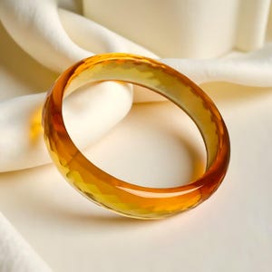May include: A faceted amber-coloured bangle bracelet. The transparent bracelet has a warm, golden hue and is set against a soft, off-white background. The bangle's surface is textured with geometric cuts, catching the light.