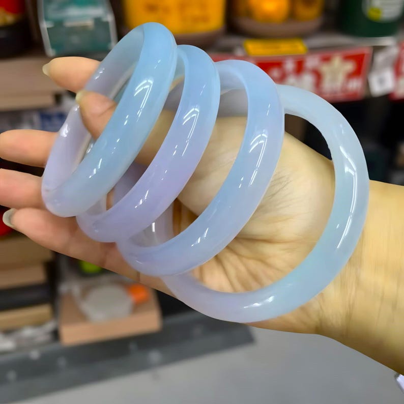 May include: Four translucent, light blue and lavender jade bangles are held in a hand. The smooth, polished bracelets have a rounded shape and varying shades of color. The background is blurred, suggesting a store setting.