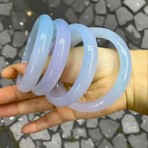 May include: Four translucent, light blue and lavender bangles are held in a hand. The smooth, rounded bracelets have a glossy finish. The bangles are stacked together, showcasing their varying shades of color.