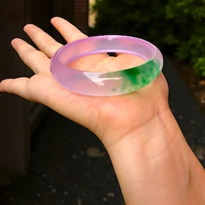 May include: A translucent jade bangle bracelet. The bracelet is a mix of colors, with a gradient of light purple to green. The green section has darker green spots. The bracelet is smooth and polished, reflecting light.