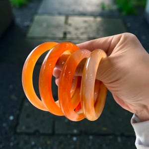 Peut inclure: Quatre bracelets-joncs orange translucides sont tenus dans une main. Les bracelets ont une surface lisse et polie et varient légèrement en couleur, allant d'un orange vif à un pêche pâle. Les bracelets sont empilés.