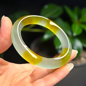 Könnte beinhalten: Ein transluzentes Jade-Armreif-Armband mit einer Mischung aus hellgrünen und gelben Farbtönen. Die polierte Oberfläche des Armbands reflektiert das Licht und hebt die natürlichen Farbvariationen hervor. Das Armband wird in einer Hand gehalten, wodurch seine kreisförmige Form und sein elegantes Design zur Geltung kommen.
