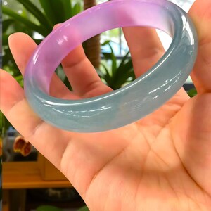 May include: A translucent jade bangle bracelet. The bracelet transitions from a soft pink hue to a pale green. The smooth, polished surface reflects light, showcasing the natural variations in color and texture. A piece of jewelry.