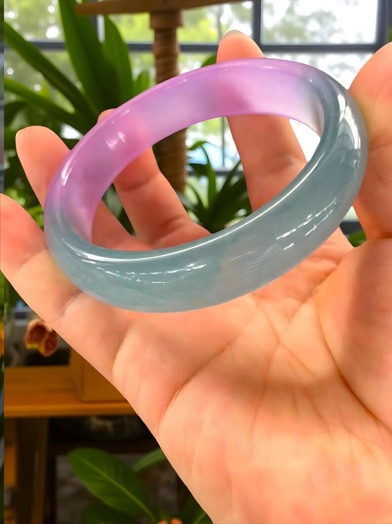 May include: A translucent jade bangle bracelet. The bracelet transitions from a soft pink hue to a pale green. The smooth, polished surface reflects light, showcasing the natural variations in color and texture. A piece of jewelry.