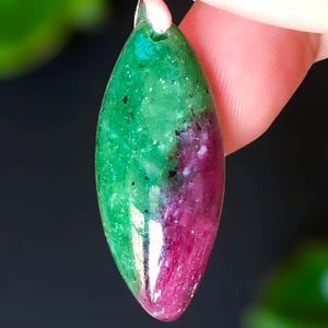 Puede incluir: Un colgante ovalado pulido con una llamativa división de color. La mitad superior es verde moteado, que se transforma en un magenta intenso en la parte inferior. Una argolla plateada está fijada en la parte superior, lo que sugiere que es una joya.