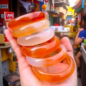 May include: A hand displays a stack of translucent, orange and white gemstone bangles. The bangles have a smooth, polished surface and vary in shades from a deep orange to a pale white. The bracelets are stacked in a cascading arrangement.