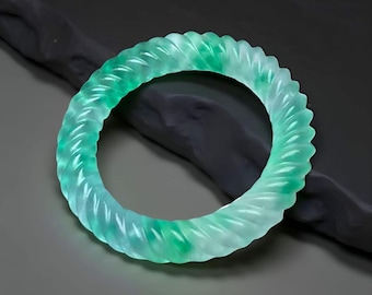 May include: A translucent jade bangle bracelet with a twisted rope design. The bracelet features a gradient of green and white hues, with a polished finish. The jewelry is displayed on a dark gray surface.