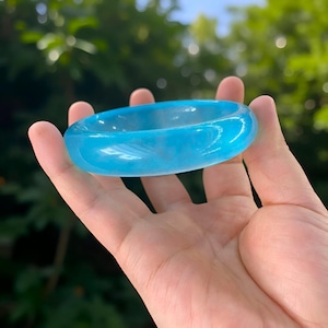 May include: A translucent, light blue bangle bracelet. The smooth, rounded bracelet is held in a hand, with a blurred green background. The bracelet's color is a vibrant, clear blue, suggesting it may be made of glass or a similar material.