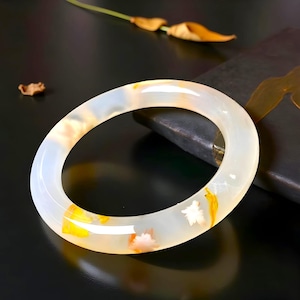 May include: A translucent jade bangle bracelet with white, yellow, and orange inclusions. The smooth, polished surface of the bracelet reflects light, showcasing the natural patterns within the stone. This elegant piece is a beautiful accessory.
