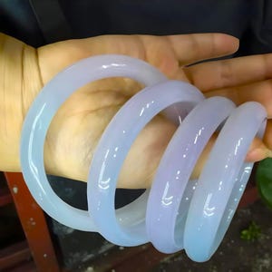 May include: Four light blue jade bangles are displayed. The translucent bracelets have a smooth, polished surface. The bangles are stacked together, creating a spiral effect. The color transitions from a lighter blue to a slightly purplish hue.