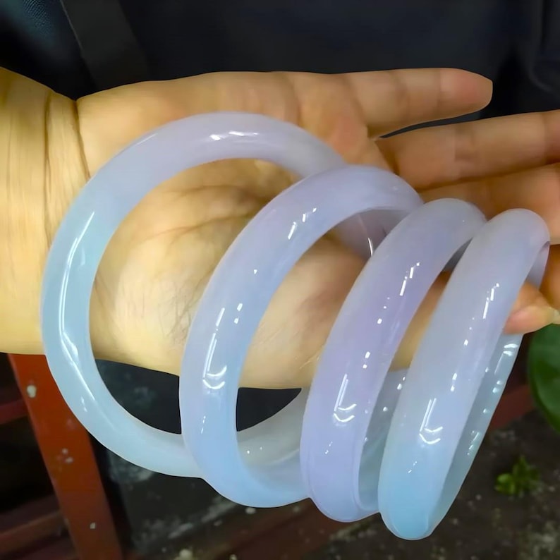 May include: Four light blue jade bangles are displayed. The translucent bracelets have a smooth, polished surface. The bangles are stacked together, creating a spiral effect. The color transitions from a lighter blue to a slightly purplish hue.