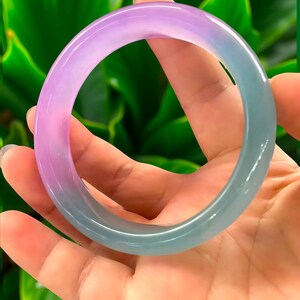 May include: A translucent jade bangle bracelet with a gradient of colors, transitioning from lavender to teal. The smooth, polished surface reflects light, showcasing the natural variations in the stone. The bracelet is held against a backdrop of green foliage.