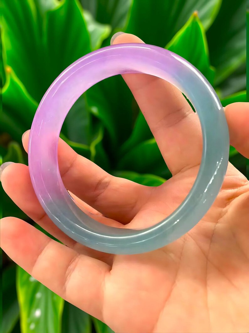 May include: A translucent jade bangle bracelet with a gradient of colors, transitioning from lavender to teal. The smooth, polished surface reflects light, showcasing the natural variations in the stone. The bracelet is held against a backdrop of green foliage.