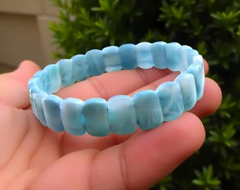 Puede incluir: Una pulsera de piedras preciosas azul claro. La pulsera está hecha de piedras rectangulares pulidas con apariencia veteada. Las piedras están ensartadas para formar una banda flexible. La pulsera se sostiene en una mano sobre un fondo de follaje verde.