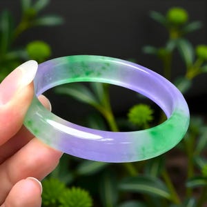 Può includere: Un braccialetto bangle in giada traslucida con una sfumatura di tonalità verdi e viola. La superficie liscia e lucida riflette la luce, mettendo in risalto le variazioni naturali di colore. Il braccialetto è tenuto su uno sfondo verde sfocato.