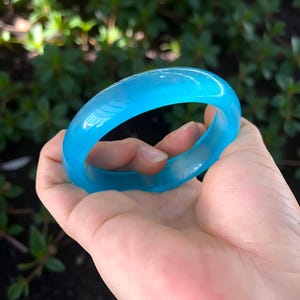 May include: A translucent, light blue bangle bracelet. The smooth, rounded bracelet is held in a hand against a blurred green background. The bangle is a solid, circular shape, and the light passes through it, giving it a glowing appearance.