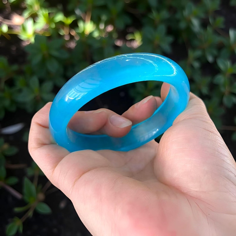 May include: A translucent, light blue bangle bracelet. The smooth, rounded bracelet is held in a hand against a blurred green background. The bangle is a solid, circular shape, and the light passes through it, giving it a glowing appearance.
