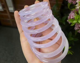 Peut inclure: Une pile de bracelets translucides de couleur lavande. Les bracelets sont lisses et arrondis, avec un éclat subtil. La teinte violet clair est uniforme sur chaque pièce. Les bracelets sont empilés dans une main, avec un arrière-plan floral flou.