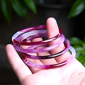 May include: Several translucent purple and red bangles. The bracelets are held in a hand, with the colours blending and swirling. The bangles are made of a smooth, polished material, and the light catches the colours.