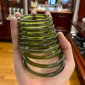 May include: A stack of olive green bangles. The bracelets are smooth and circular, with a translucent quality. The bangles are held in a hand, with a blurred background of a display case.