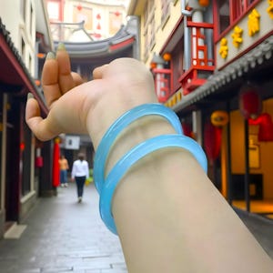 Puede incluir: Dos brazaletes azul claro se llevan en la muñeca. Los brazaletes son de un color sólido translúcido. El fondo es una escena borrosa al aire libre con edificios y personas.