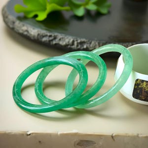 May include: Three translucent green jade bangles. The bracelets are oval-shaped and have a smooth, polished surface. The bangles are stacked together, with a white teacup and a dark gray plate in the background.