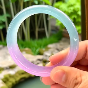 May include: A translucent jade bangle bracelet with a gradient of colors, transitioning from a light blue to a soft purple. The smooth, polished surface reflects light, showcasing the natural variations in the stone. The bracelet is held against a blurred green background.