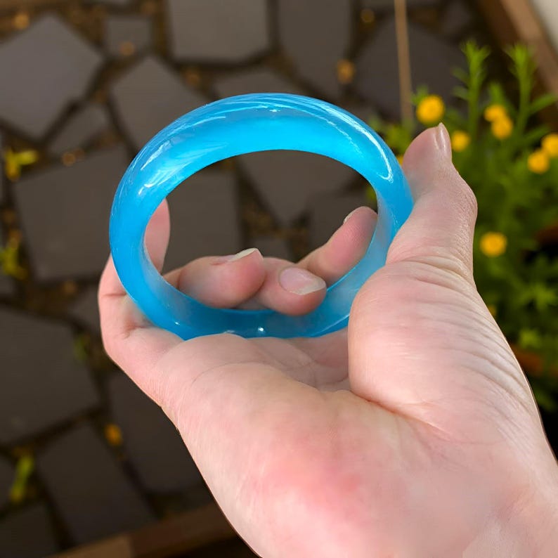May include: A translucent, light blue bangle bracelet. The smooth, rounded bracelet is held in a hand, showcasing its vibrant color and simple design. The bangle is a solid, circular shape, perfect for everyday wear.