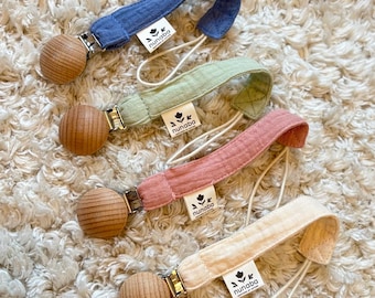 May include: Four baby pacifier clips in blue, green, pink, and cream. Each clip has a wooden bead, a fabric loop, and a metal clasp. The fabric loops have the brand name "nunaba" printed on them.