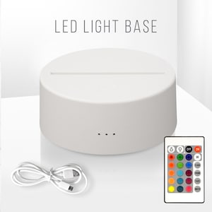 May include: A white LED light base with a slot for displaying an object. The base has three small holes and includes a remote control with color options. A white USB cable is also included. The text "LED LIGHT BASE" is at the top.