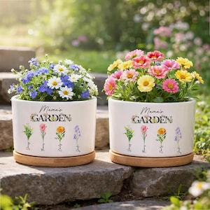 May include: Two white ceramic flower pots with the text "Mama's Garden" and floral illustrations. One pot contains blue and white flowers, the other has pink, yellow, and red flowers. Both pots sit on wooden saucers.