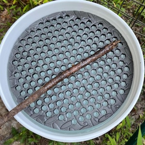 Könnte beinhalten: Ein weißer Plastikeimer mit einem grauen Einsatz mit Wabenmuster. Ein brauner Stock liegt diagonal über dem Einsatz. Der Eimer befindet sich im Freien, mit grünem Laub im Hintergrund.